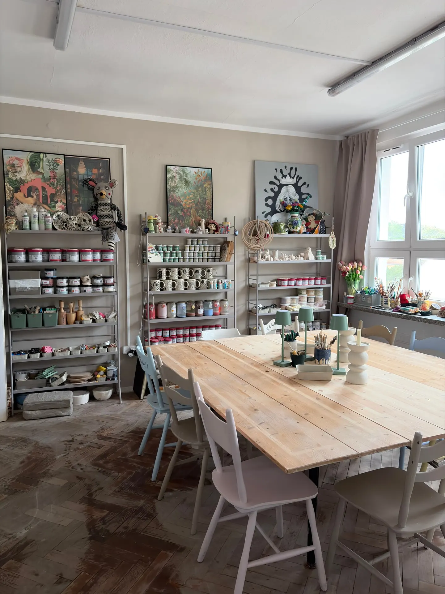 Długi drewniany stół w pracowni, pastelowe krzesła i regały pełne farb ceramicznych